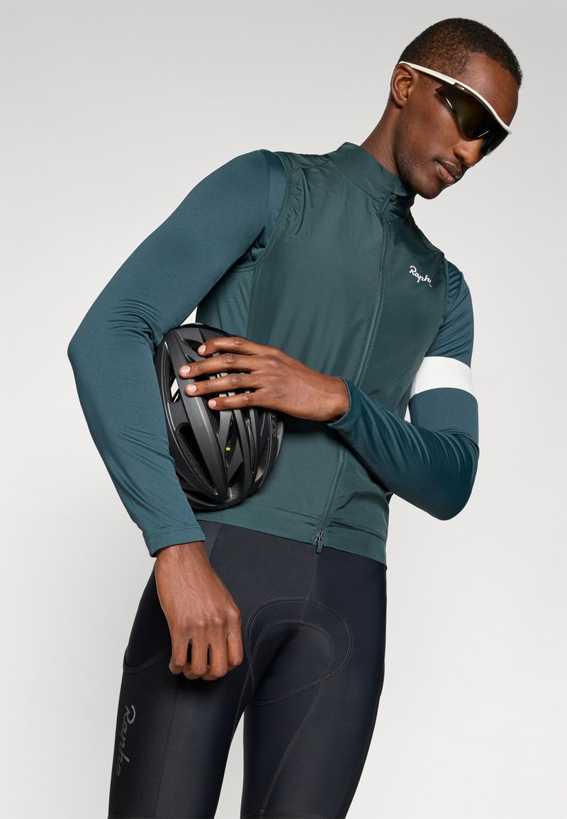 Tenue de cyclisme comprenant un gilet vert foncé et un short rembourré noir, avec un casque noir et des lunettes de soleil, mettant en avant un design et un ajustement athlétiques modernes.