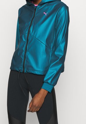 Personne portant un sweat à capuche zippé bleu sarcelle brillant avec un logo Puma rose et des leggings noirs, debout devant un fond clair uni.