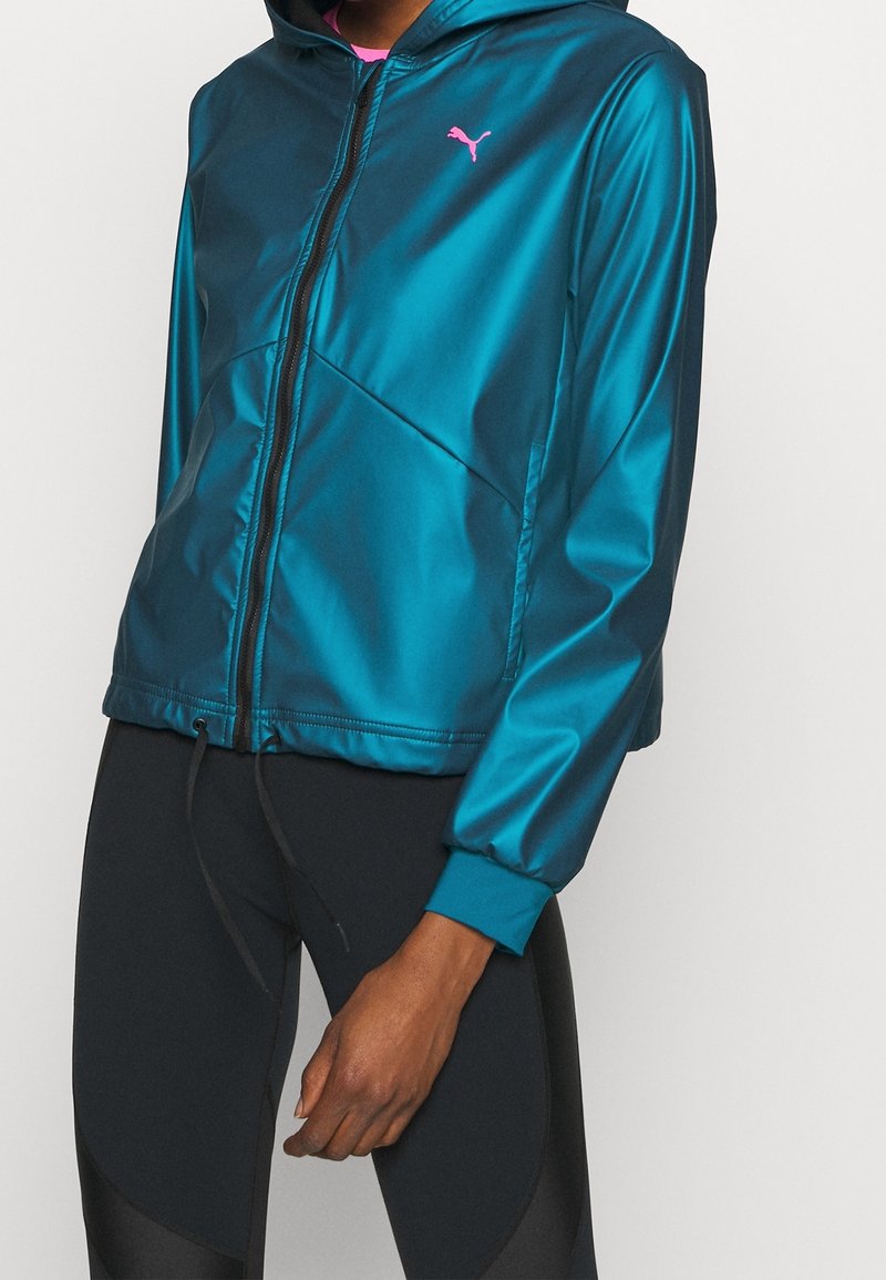 Personne portant un sweat à capuche zippé bleu sarcelle brillant avec un logo Puma rose et des leggings noirs, debout devant un fond clair uni.