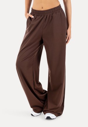 Kobieta nosząca luźne, brązowe spodnie z szerokimi nogawkami, z elastycznym pasem i białe trampki, stoi z jedną ręką w kieszeni.