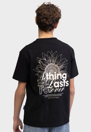 Persoon met kort krullend haar draagt een zwart T-shirt met een grote zonnebloem en witte tekst "Nothing Lasts Forever" op de achterkant.