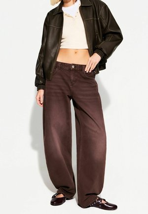 Frau trägt braune Jeans mit weitem Bein, beiges cropped Top, dunkelbraune Lederjacke und dunkelbraune flache Schuhe mit Schnürsenkeln, steht vor weißem Hintergrund.