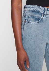 Primer plano de la mano izquierda de una persona descansando al lado de unos jeans de mezclilla azul claro con el bolsillo delantero y los pasadores del cinturón visibles.