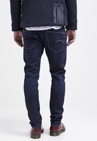 Jeans skinny blu scuro con una leggera lucentezza, tasche posteriori cucite, abbinati a un maglione di lana a trama navale e scarpe marroni.