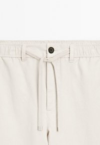 Pantaloni di cotone beige chiaro con vita elastica, coulisse e un singolo bottone nero sul davanti. Sono visibili tasche anteriori piatte.