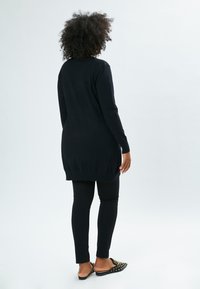 Schwarze langärmelige Strickjacke mit lockerem Schnitt, geribbtem Saum, kombiniert mit schwarzen Leggings und verzierten Slippern. Einfache, glatte Textur.