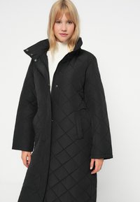 Juodas gumuotas ilgas paltas su aukšta apykakle, uždarytas spaudiniais mygtukais, su šoninėmis kišenėmis ir lygiomis tekstūra. Dėvimas ant kreminės spalvos megztinio.