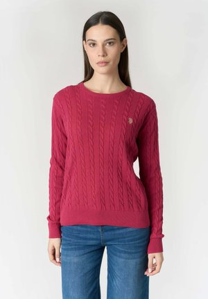 Donna con maglione rosa a coste a maniche lunghe e jeans blu, in piedi davanti a uno sfondo chiaro e semplice.