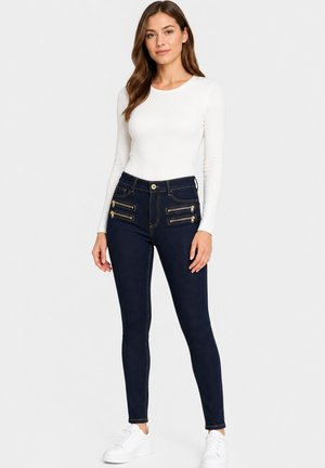 Vrouw draagt een witte top met lange mouwen, donkerblauwe skinny jeans met gouden ritsdetails en witte sneakers, staand voor een effen achtergrond.
