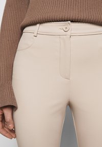 Pantalons beiges taille haute en tissu lisse, dotés d'une fermeture à un bouton et de poches latérales. La taille est soigneusement taillée.
