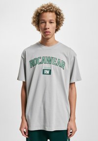 Harmaa puuvillatrikoopaita, jossa on vihreä "ROCAWEAR" -logo ja teksti "EST. 1999". Lyhyet hihat ja rento istuvuus.