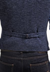 Marinblå stickad väst med en texturerad yta, som har ett litet justerbart bälte på baksidan och kontrasterande tyg som tittar fram under.