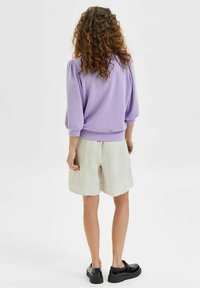Suéter lila con mangas abullonadas, combinado con pantalones cortos crema. La modelo lleva calzado negro, mostrando una textura suave y un diseño casual.