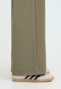 Olivengrønne, brede bukser med en glat tekstur, parret med beige sneakers, der har tre sorte striber og en let tekstureret sål.