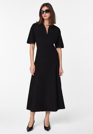 Schwarzes Midikleid mit kurzen Ärmeln, V-Ausschnitt und zwei Brusttaschen. Glatte Textur und ausgestellter Rock. Kombiniert mit schwarzen spitzen Absatzschuhen.