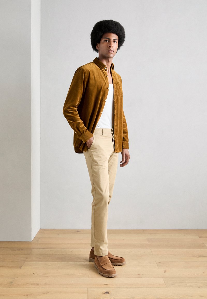 Braunes Cordhemd mit Knöpfen, weißes Tanktop, beige Hose und braune Wildleder-Loafers. Das Model steht vor einem schlichten Hintergrund.