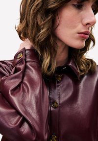 Bordeauxrode leren jas met een glanzende afwerking, voorzien van een kraag, grote knopen en een geplooid ballonmouwontwerp.