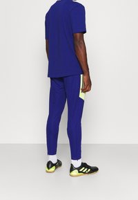 Mężczyzna w niebieskich spodniach sportowych z żółtymi panelami kieszeni, dopasowanej niebieskiej koszulce, białych skarpetkach i czarnych butach sportowych do użytku wewnętrznego, stojący przy białej ścianie.