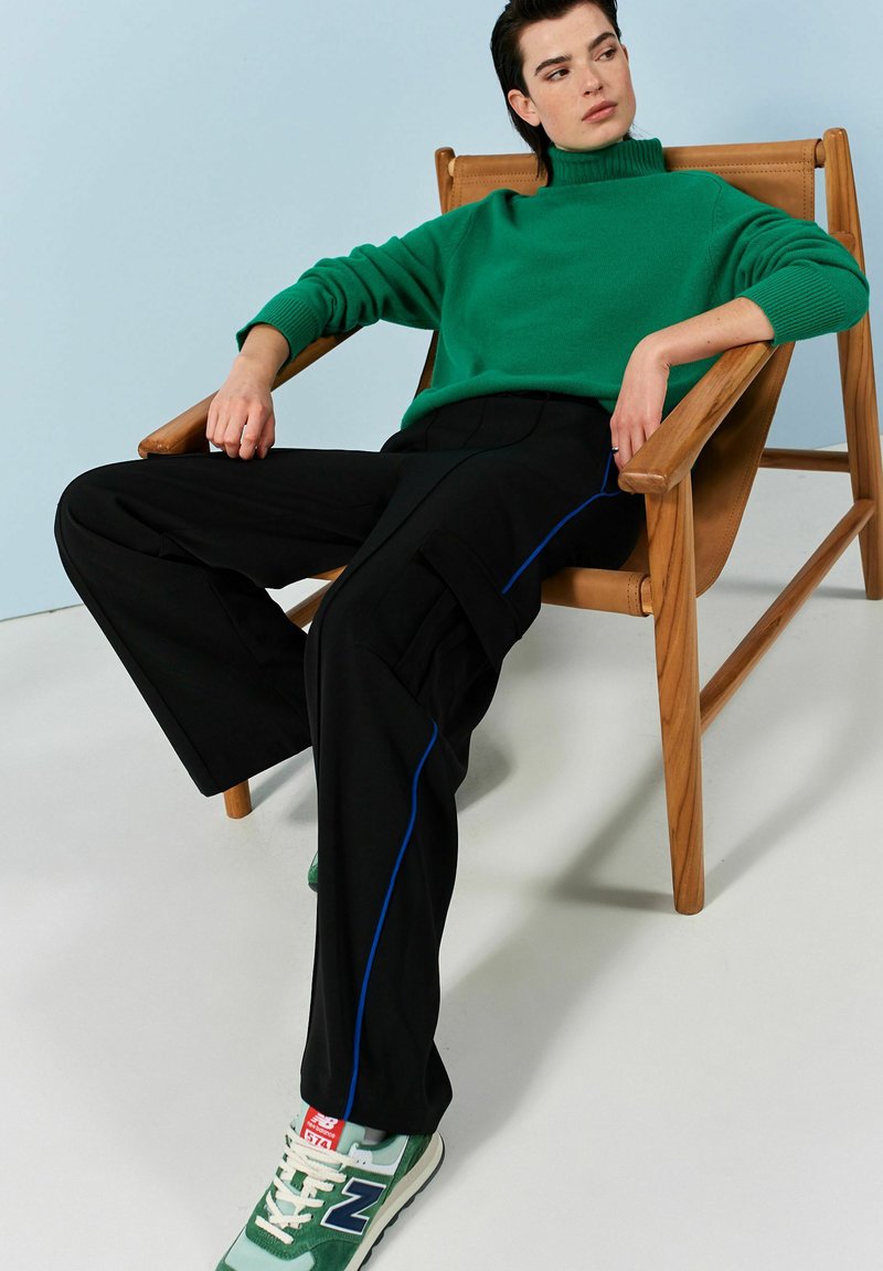 Grön stickad polotröja, svarta vida byxor med blå passpoal och gröna New Balance-sneakers på en trästol mot en ljus bakgrund.