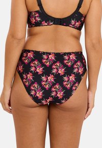 Femme portant des culottes noires à fleurs taille haute et un soutien-gorge assorti, vue de dos sur fond blanc.