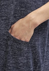 Stickad, mörkgrå tyg med en mjuk textur. Synlig ficka på vänster sida, med handen inskjuten, vilket visar plaggets avslappnade design.