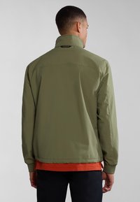 Giacca leggera di colore verde oliva con colletto alto, polsini elasticizzati e vestibilità rilassata. Presenta un sottile dettaglio logo vicino al colletto.