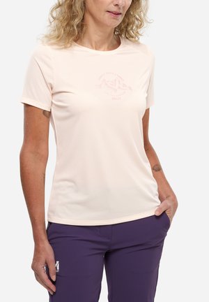 Femme portant un t-shirt rose clair à manches courtes avec un logo montagne et un pantalon violet, une main dans la poche, sur fond blanc.