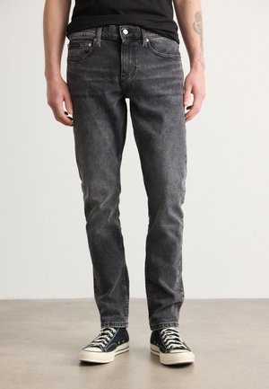 Personne portant un jean slim noir délavé et des baskets montantes noires, debout sur un sol en béton lisse contre un mur clair uni.