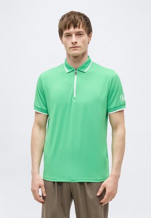 Bărbat tânăr purtând un tricou polo cu mânecă scurtă de culoare verde aprins, cu fermoar alb și logo pe mâneca stângă, stând în fața unui fundal simplu.