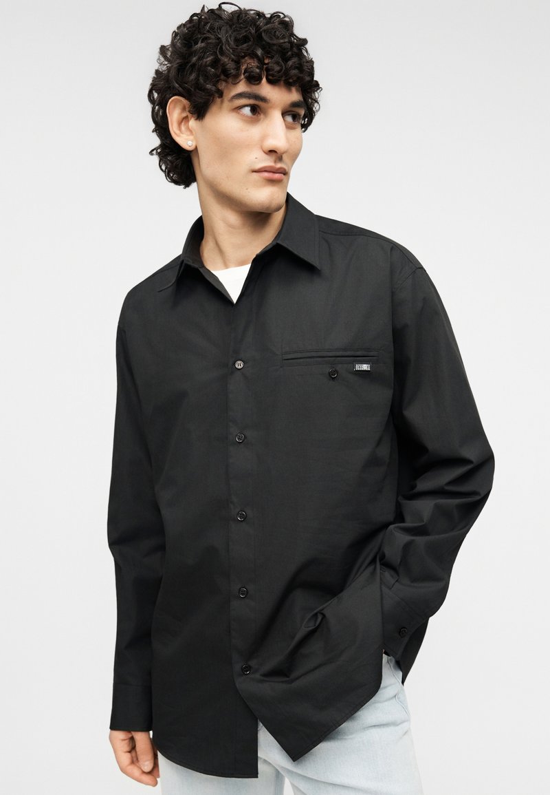 Chemise noire à boutons avec manches longues, dotée d'une poche poitrine et d'une étiquette de marque discrète. Texture lisse, col standard, coupe ajustée.