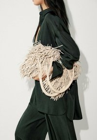 Beige hapsulaukku, jossa on teksturoidusta materiaalista tehdyt, kaarevat kahvat ja kerroksellinen muotoilu, pidetty tummanvihreää asua vasten.