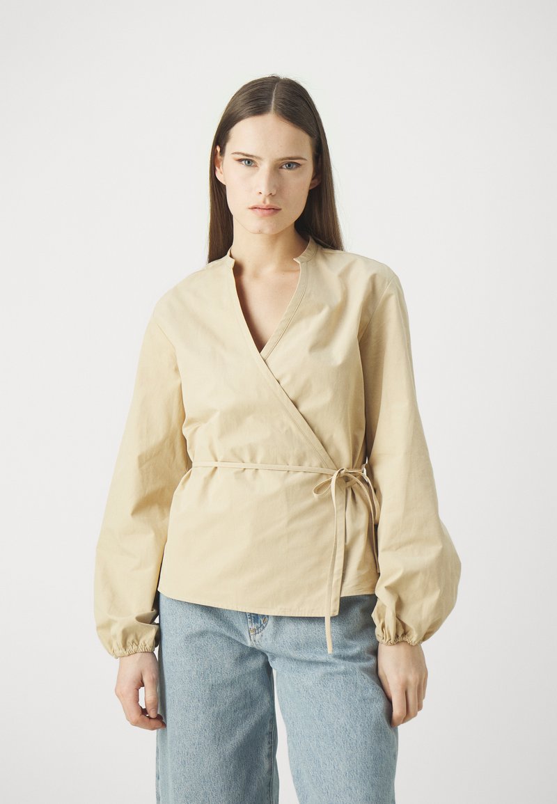 Beige wikkelblouse van katoen met een V-hals, lange pofmouwen en een verstelbare strik in de taille, gecombineerd met lichtblauwe jeans.