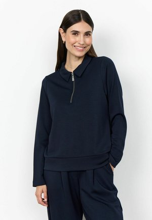 Žena s dlhými tmavými vlasmi sa usmieva, má na sebe tmavomodrý top s dlhými rukávmi a predným zipsom a tmavomodré nohavice, ruka v vrecku.