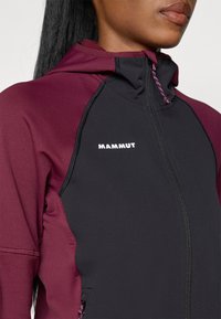Casaco ajustado preto e bordô com capuz, apresentando um fecho frontal e o nome da marca "Mammut" impresso em branco. Têxtil texturizado com costura em contraste.
