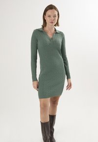 Robe en tricot de couleur vert sourd avec un motif en relief, col V et manches longues. Comprend un plastron boutonné et des poignets et un ourlet côtelés.