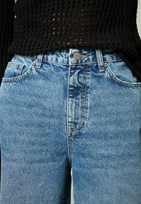 Jean en denim bleu avec une taille haute, un design classique à cinq poches et des coutures contrastées. Le matériau présente une texture légèrement décolorée.