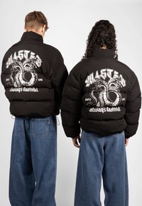 Black puffer jacket with graphic design featuring snakes and text on the back, paired with blue denim wide-leg pants.