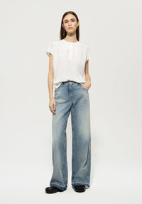 Weißes kurzärmliges Oberteil mit gerafftem Ausschnitt. Hellblaue weite Jeans mit verwaschenem Look. Schwarze Plateausandalen. Neutraler Hintergrund.