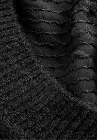 Tissu noir texturé avec un côté tricoté duveteux et une section lisse et transparente présentant des accents de fils horizontaux et de petits fermoirs circulaires.