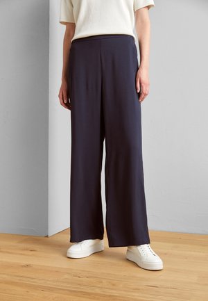 Person, die lockere, hoch taillierte schwarze Hose mit weitem Bein und weiße Turnschuhe trägt, steht auf einem hellen Holzboden vor einer grauen Wand.