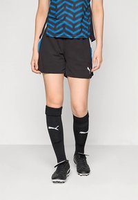 Melnas šorti ar zilām sānu akcentiem, kombinēti ar melnām ceļgalu garuma zeķēm ar baltu logotipu un melnām futbola kedām.