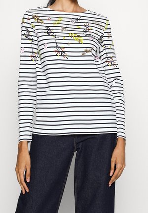 Woman wearing a white long-sleeve top with black stripes and floral print, paired with dark blue jeans.