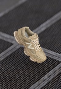 Chaussure de sport beige avec des matériaux en nylon et en daim, dessus texturé, bout arrondi, semelle épaisse et logo bien visible. Lacets de couleur assortie.