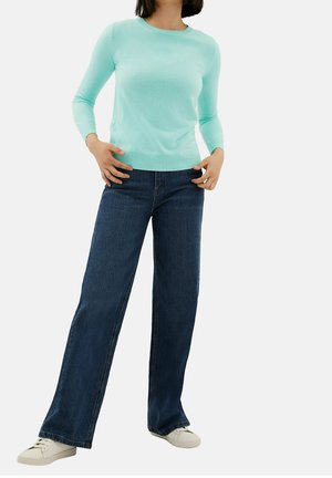 Femme portant un pull à manches longues bleu clair, un jean à jambes larges bleu foncé et des baskets blanches, debout les mains près des hanches sur fond blanc.