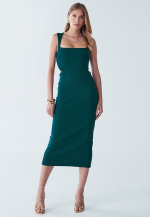 Teal fitted dress with square neckline and cut-out sides. Made of textured fabric, it falls to mid-calf and is paired with strappy sandals.