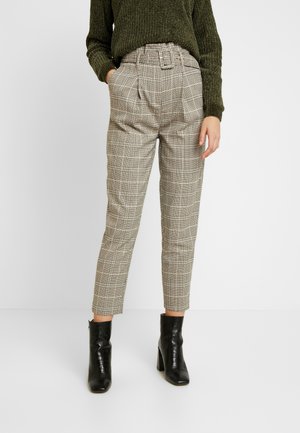 Kobieta w beżowych w kratę spodniach z wysokim stanem i paskiem, zielonym dzianinowym swetrze oraz czarnych botkach na obcasie, stojąca na tle białego tła.