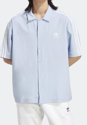 Personne portant une chemise à manches courtes bleu clair avec boutonnière Adidas et des rayures blanches sur les manches, ainsi qu'un pantalon blanc avec le logo Adidas.