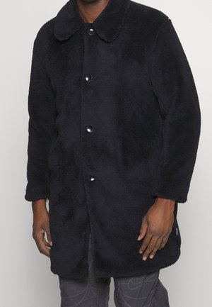 Manteau en polaire noir à boutons avec col, porté par une personne dont les mains sont visibles, associé à un pantalon sombre à motifs.