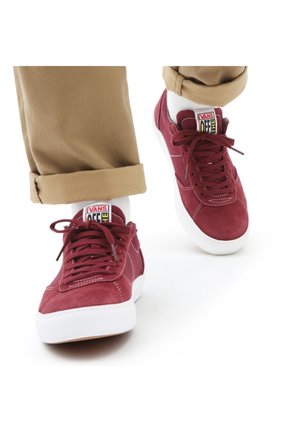 Vans tenisky z tmavě červené semišové kůže s bílými gumovými podrážkami. Mají ploché tkaničky, polstrované límce a barevné logo na jazyku. Viditelné jsou zavinuté khaki kalhoty.