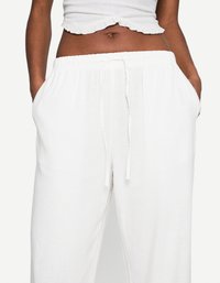 Modèle portant un crop top léger à volants et un pantalon blanc à cordon de serrage, les mains dans les poches sur un fond uni.